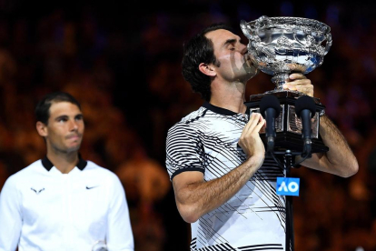 Roger Federer besa el trofeo conseguido en Melbourne con Rafa Nadal al fondo de la imagen.