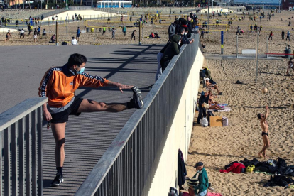 Un joven con mascarilla hace estiramientos tras hacer ejercicio en una playa barcelonesa.