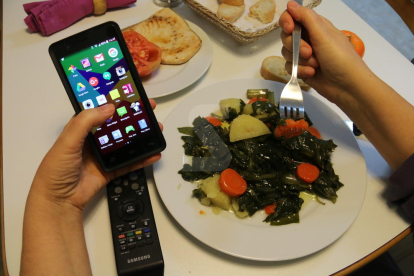 Un menor menja amb un mòbil en una mà.
