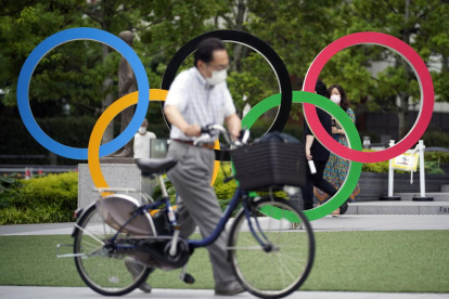 Un ciutadà de Tòquio camina amb la bicicleta davant d’uns grans anells olímpics.