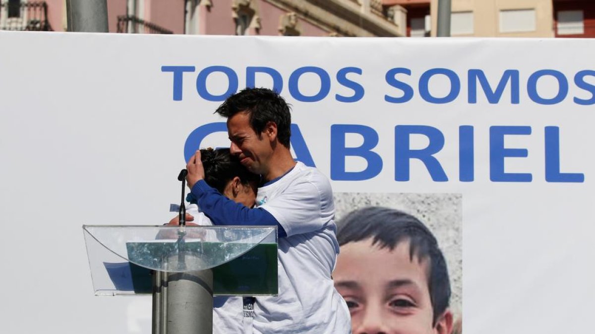 Los padres de Gabriel, abrazándose delante de una fotografía del menor en un acto el pasado viernes y, a la derecha, la detenida, Ana Julia Quezada.