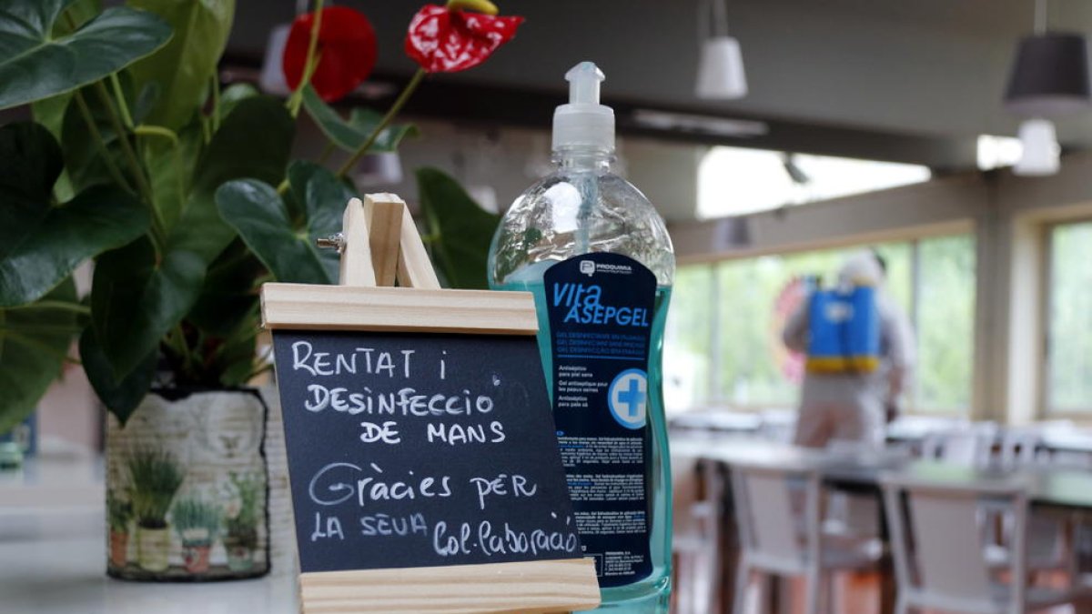 Un cartel y una botella de gel desinfectante, mientras en el fondo un trabajador del restaurante El Gat de Tàrrega desinfecta el local.