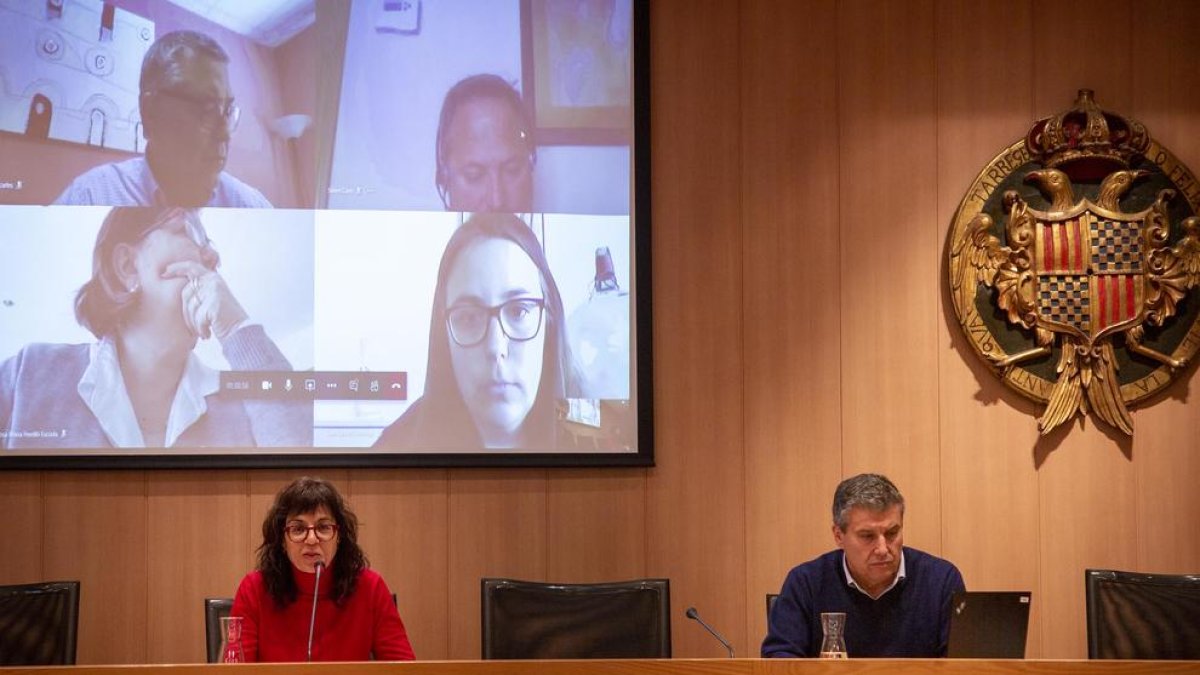 L’alcaldessa i el secretari municipal a la sala de plens. A la pantalla, alguns regidors.