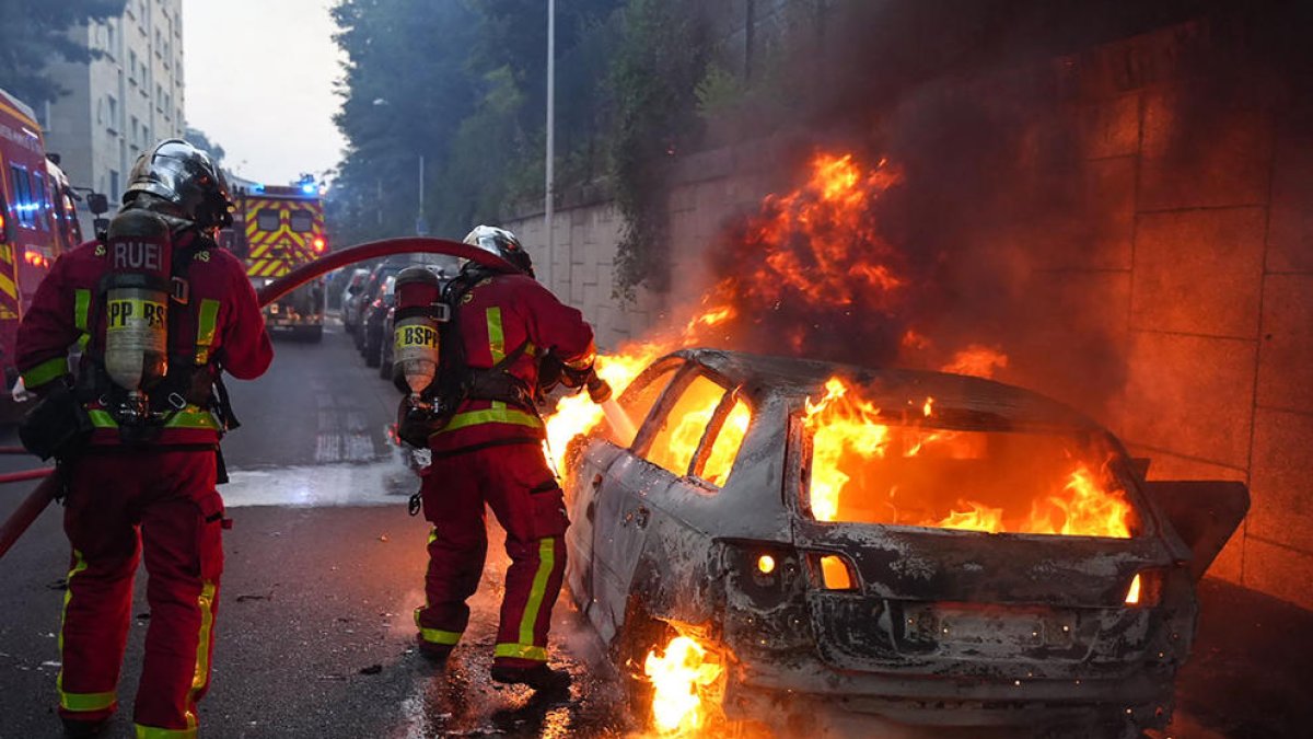 Els bombers apaguen les flames d’un cotxe durant els disturbis.