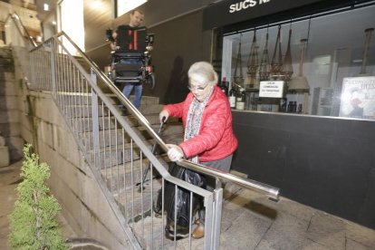 Regina baja las escaleras de espaldas mientras un vecino le lleva la silla de ruedas para entrar en casa.