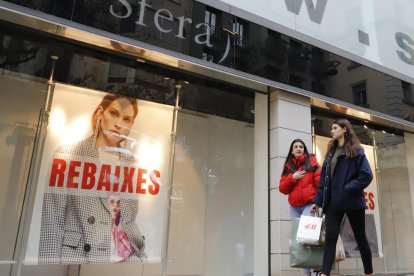 Dos jóvenes con bolsas pasan delante de un cartel de rebajas de una tienda del Eix Comercial. 