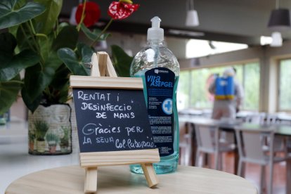 Un cartel y una botella de gel desinfectante, mientras en el fondo un trabajador del restaurante El Gat de Tàrrega desinfecta el local.