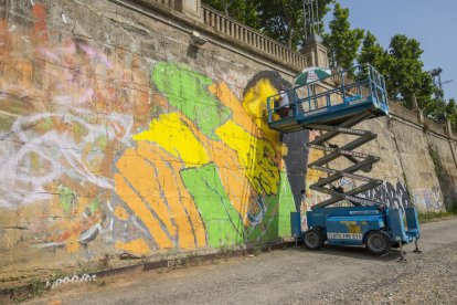 L’artista Llukutter pintant el mural ‘Lleida ciutat acollidora’ al mur de la canalització del riu.