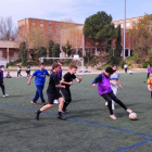 Un grupo de alumnos juega un partido de fútbol en el Camp Escolar con el instituto Joan Oró al fondo. 