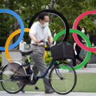 Un ciutadà de Tòquio camina amb la bicicleta davant d’uns grans anells olímpics.
