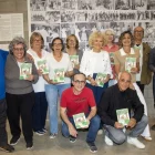 La junta dels jubilats amb l’alcaldessa mostrant el llibre de receptes davant d’un plafó. - LAIA PEDRÓS