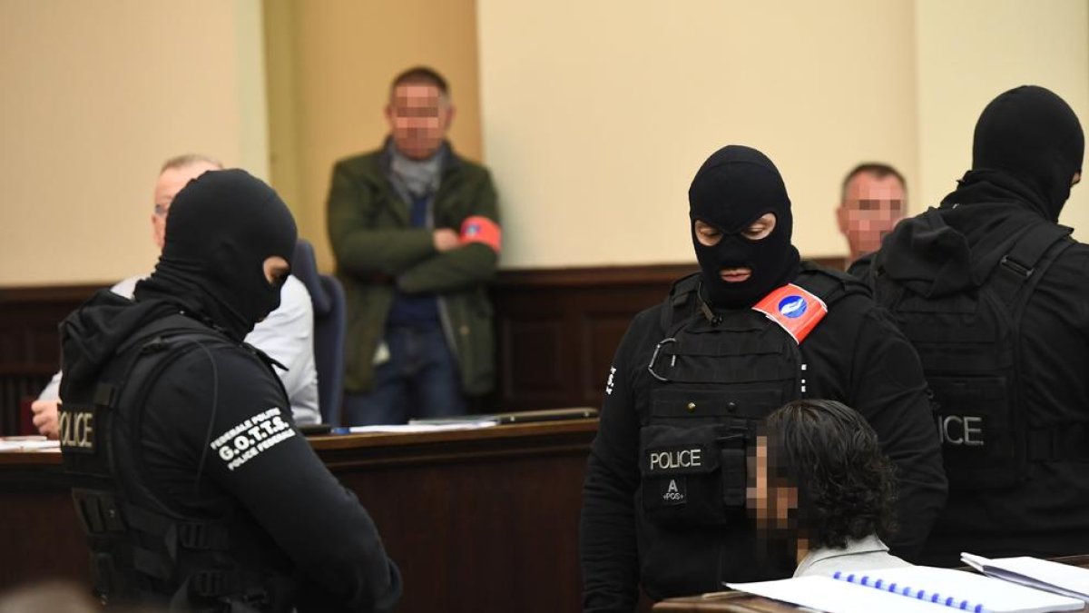 Agentes de policía enmascarados custodian en el tribunal a Salah Abdeslam, cuya cara está pixelada.