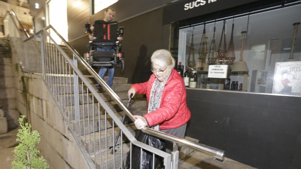 Regina baixa les escales d’esquena mentre un veí li porta la cadira de rodes per entrar a casa.