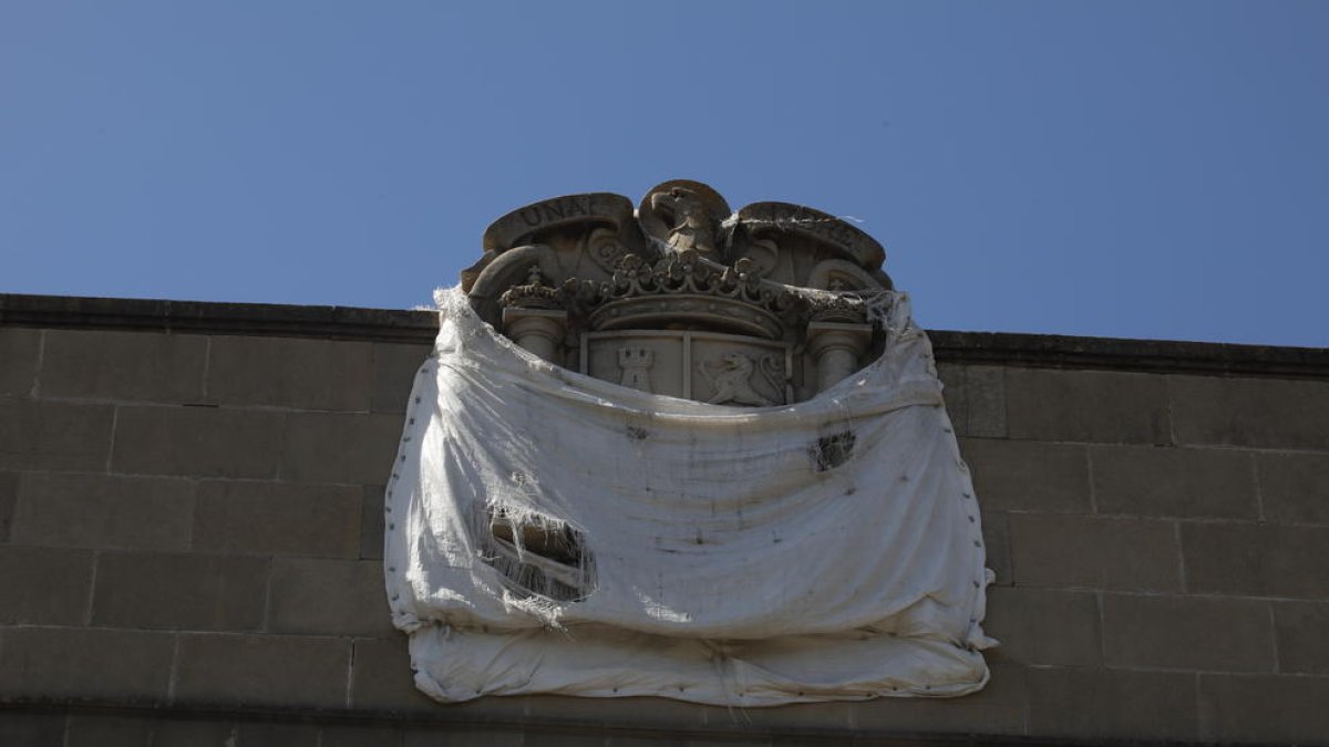 Mig tapat - L’escut amb l’àguila franquista esculpit en pedra a dalt de l’edifici porta anys mig tapat amb una lona blanca, que periòdicament es despenja i el deixa de nou al descobert, tot o en part. Actualment, presenta aquest aspecte, mi ...