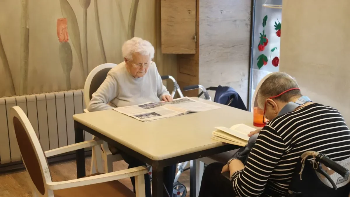 Una mujer lee el diario en una residencia junto a otro residente con un libro. - ACN